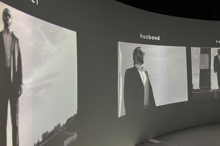 Black and white projected image of a man standing in a sit with text that says husband. From the Cradle of Fire film by Diana Scarborough