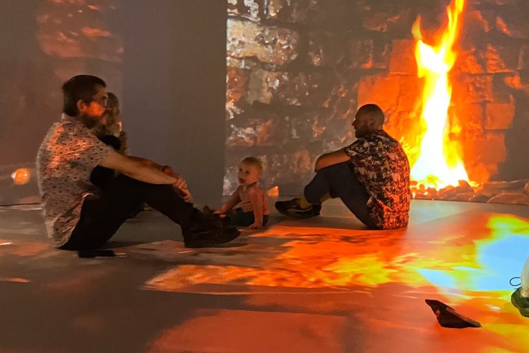 adults in babysitting on teh floor against a projection screen showing bricks and flames from a forge